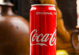 Na fotografiji je crvena limenka Coca‑Cole s bijelim logotipom i natpisom "ORIGINAL TASTE" na vrhu. Pozadina je zamućena, s tamnim i smeđim tonovima.